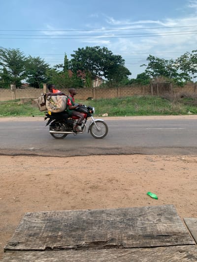 The image depicts a scene in Kabba, Nigeria. A motorcycle with two passengers is traveling along a paved road. The passengers include a driver and a passenger sitting behind him. The passenger is carrying what appears to be a bag or luggage wrapped in a fabric. The roadside consists of a dirt area with some scattered debris, including a green plastic wrapper, and a weathered wooden structure on the right side of the image. On the left of the road is grass, trees, and a brick wall. Overhead power lines are visible in the background against a cloudy sky.