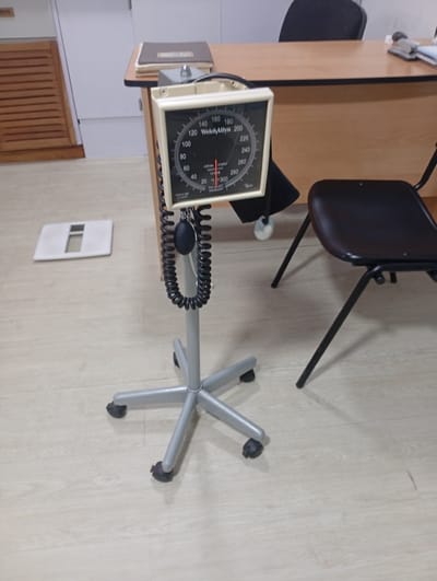 The image shows a medical examination room. In the foreground, a portable aneroid sphygmomanometer on a wheeled stand is the primary subject. The device has a black dial with white markings indicating blood pressure in mmHg, ranging from 20 to 300. A black bulb and coiled tubing are attached to the device. To the left of the sphygmomanometer, a white, flat electronic scale with a cutout is visible on the light-colored wooden floor. In the background, a wooden desk with a stapler and some papers is present. Behind the desk, a black office chair is partially visible. The room appears to be part of a medical facility, possibly a clinic or doctor's office, and is located in Valencia, Venezuela.