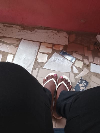 The image provides a top-down view of an individual's lower legs and feet in Isasi, Nigeria. The person is wearing black trousers and white-colored flip-flops. Their dark-skinned feet exhibit toenails painted a reddish-pink hue. The feet are positioned on an uneven ground surface constructed from numerous fragmented and irregularly shaped ceramic tiles. These tile pieces vary in color, including white, beige, and dark blue with a floral pattern, and are set into a coarse, possibly concrete or earthy, substrate. Behind this tiled floor, a reddish-pink wall with a lighter, unpainted concrete base is visible.