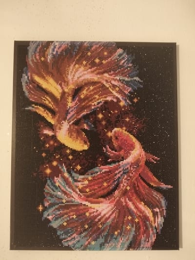 This is a diamond painting of two betta fish in a celestial setting. The fish are depicted in profile, facing each other, and appear to be swimming in a galaxy or starry night sky. One fish is predominantly yellow and orange, while the other is red and pink, with accents of blue and purple in their flowing fins. Small, sparkling diamond-like beads are scattered throughout the dark background, representing stars. The overall impression is one of movement and vibrant color against a dark, ethereal backdrop. The diamond painting is framed in black and mounted against a plain, light-colored wall. There is no visible text in the image. The location context is Қараөткел, Kazakhstan.