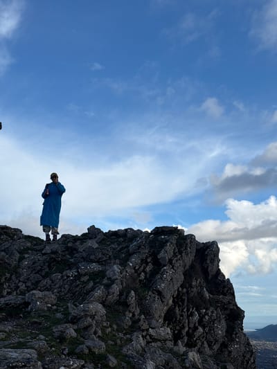 An individual stands atop a rugged, rocky mountain peak in Bogdašići, Montenegro. The person is positioned in the central-left of the frame, facing slightly to their left. They wear a long, loose-fitting, light blue coat, light-colored pants, and dark boots. A beige baseball cap covers their head, and dark sunglasses obscure their eyes. In their right hand, they hold a black smartphone, seemingly held up for a photograph or video. A red-colored cylindrical object, possibly a can or bottle, is held in their left hand. A dark backpack is visible on their back. The ground consists of numerous dark grey, craggy rock formations with sparse patches of green vegetation. The sky above is largely clear blue with scattered white and grey cumulus clouds. In the far background, beyond the rocky precipice, a body of water (likely the Adriatic Sea) and a coastal settlement with structures are visible, nestled against distant mountains.