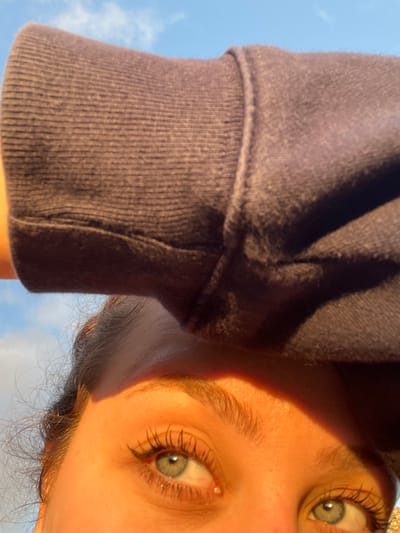 The image shows a person's face at an unusual angle, with the sky visible in the background. The person has fair skin, light-colored eyes, and visible eyelashes. A portion of a gray-purple sweatshirt is also visible, suggesting the person is wearing it. The photo appears to be taken outdoors, possibly in the daytime. There is no information to suggest that this photograph was taken in Mogán, Spain.