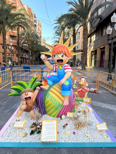 This is a vertical, medium shot of a large, colorful, street art display, likely a "falla" from the Valencian Community, Spain. The subjects are two cartoonish figures, one larger with orange hair and a yellow sunburst headdress, wearing a blue outfit with red and white striped sleeves, and holding a pocket watch. The other figure is smaller, lying on a bed of white pebbles, with brown hair and a shell-like headdress, wearing a brown outfit. The display is on a street, with buildings and palm trees in the background. There are also decorative elements like a small wooden structure with Easter eggs and flowers, and small signs with text in Spanish and Catalan. The scene appears to be set up outdoors during the daytime, with clear blue skies visible. The overall mood is festive and playful, indicated by the exaggerated expressions of the figures and the vibrant colors of the display. Signs visible include "FCARNER SANT VICEN", "EL TALLERET", and some handwritten text in Spanish and Catalan.