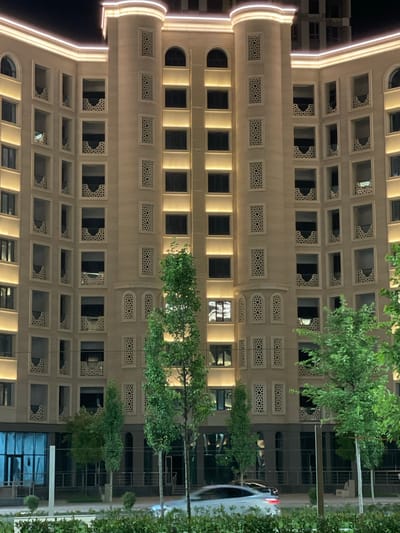 The image captures the nighttime facade of a modern, multi-story building in Grozny, Russia. The building is illuminated by warm, yellow lights that highlight its architectural features, including rows of identical windows and decorative balconies with intricate white latticework. The facade is a light beige color, and the building has rounded corners and vertical accent lighting. Trees with lush green foliage are visible in the foreground and on the sides of the building, partially obscuring the lower floors. A white car is driving past in the foreground, its headlights and taillights blurred due to motion. The scene suggests a well-lit urban environment at night.