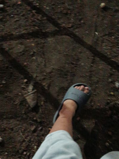 The image shows a close-up of a person's left foot wearing a gray slide sandal, walking on dark, uneven ground. The ground appears to be soil with some small stones and debris. Shadows are cast across the ground, suggesting it is outdoors, possibly during the daytime or under artificial light. The person is wearing light-colored clothing, partially visible at the bottom of the frame. The overall impression is one of a casual outdoor setting. Based on the prompt's request for location, this scene could be found anywhere within the city of Novotitarovskaya, Russia, where such ground conditions and casual footwear are common.