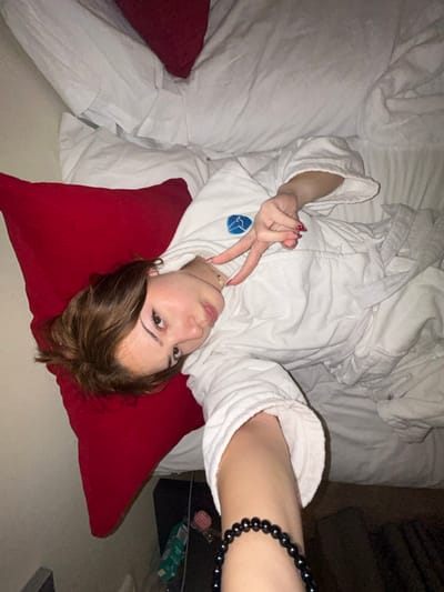 A young woman in a white bathrobe lies on a bed with white sheets, her head resting on a red pillow. She's holding up a hand making the peace sign. She is wearing a delicate gold chain necklace. A dark beaded bracelet adorns her wrist. The photo appears to be taken at an angle from above.