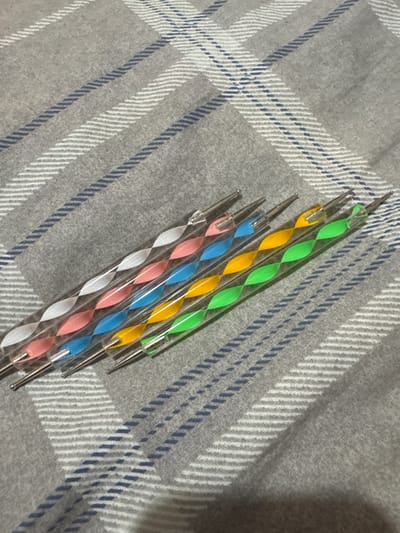 Five dotting tools with colorful spiral handles are laid out diagonally on a grey and white plaid fabric. The tools have silver metal tips. From left to right, the handles are white, pink, blue, yellow, and green. The fabric appears to be a blanket or bedspread. There is no indication of the location being in the city of Яромінскі сельскі Савет, Belarus.