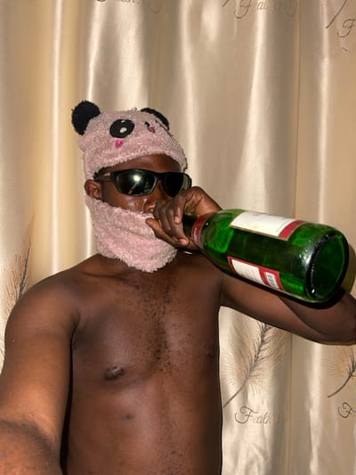 A shirtless dark-skinned person with sunglasses and a pink panda headband lies horizontally on a patterned beige surface, possibly a bed. The person is holding an upturned green glass bottle with a white label. The overall image appears unconventional and potentially humorous, given the juxtaposition of the shirtless figure with the panda headband and wine bottle.