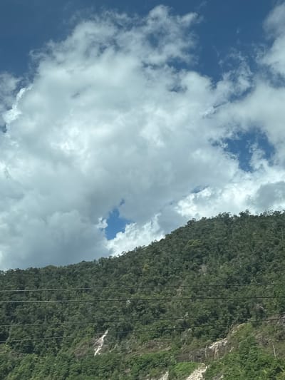The image depicts a lush, green mountainside in Nam Khanh Vinh, Vietnam. Dense forest covers the slopes, interspersed with rocky outcrops and a small, white waterfall. Several dark power lines stretch horizontally across the mid-ground of the mountain. Above the mountain, a dynamic sky is visible, filled with a mix of bright blue patches and large, billowing white clouds. The scene is a natural landscape, devoid of any visible people or human activity beyond the power lines.