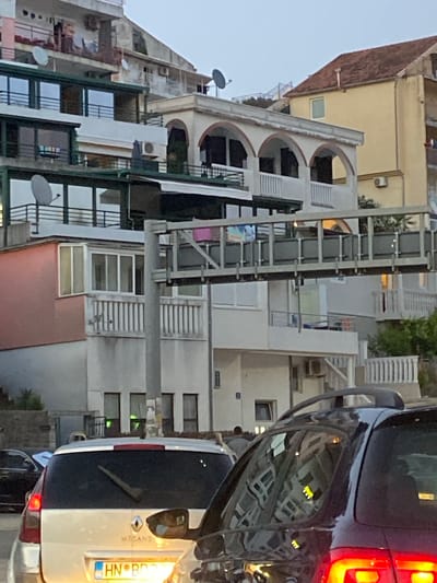 The image shows a street scene in Meljine, Montenegro, with a focus on layered buildings and vehicles. In the foreground, the rear of a light-colored vehicle, possibly a Renault Megane, is visible, facing away from the camera. Its license plate, partially illuminated, reads "HN BD...". To its right, a portion of a dark-colored vehicle is present.
Behind the cars, a multi-story building with a pink facade on its lower section and white balconies and windows on its upper levels is prominent. A covered walkway or bridge with a metal railing structure spans across the scene above the building. Further back, additional buildings with balconies, arched walkways, satellite dishes, and air conditioning units are visible. One building has a reddish-brown tiled roof. The sky is overcast. A person can be seen walking in the background between buildings. The environment suggests an urban or semi-urban residential area.