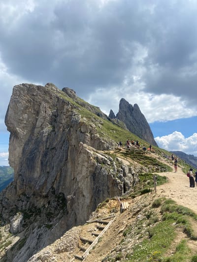 This media file captures a dramatic mountain landscape with a winding path and a group of people. The scene is characterized by steep, rocky cliffs covered in patches of green grass and wildflowers. A well-worn dirt path, bordered by a safety railing, snakes along the mountainside, indicating it is a popular hiking route. Several individuals are visible on the path, some walking, others pausing to admire the view.
The sky is filled with a mix of dramatic, dark clouds and patches of bright blue, suggesting a dynamic and possibly changing weather pattern. The time of day appears to be daytime, likely late morning or early afternoon, given the bright lighting and the shadows cast by the rocks and people.
A prominent geological feature is a large, overhanging rock formation that juts out over a steep drop, creating a sense of awe and perhaps a touch of trepidation. Wooden steps are built into the side of the mountain in some areas, providing access and support for hikers. The overall impression is one of a challenging yet rewarding outdoor adventure in a rugged, natural environment. There is no visible text in the image.
