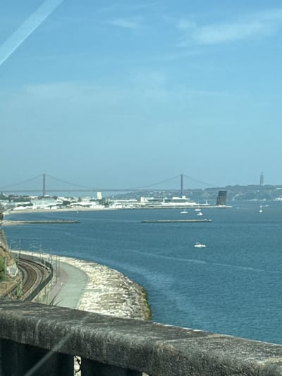 The image depicts a scenic view of the Tagus River estuary in Oeiras, Portugal, under a clear blue sky. A suspension bridge, likely the 25 de Abril Bridge, spans the river in the distance, connecting to the cityscape on the opposite bank. The cityscape features industrial buildings and a prominent dark, angular modern structure. Several boats, including a few white speedboats, are visible on the water.
In the foreground, a curved railway track runs along the edge of a rocky embankment. A paved path runs parallel to the tracks, separated by a stone wall. A textured stone railing is in the immediate foreground, framing the view. The overall impression is one of a coastal urban landscape with natural and man-made elements.