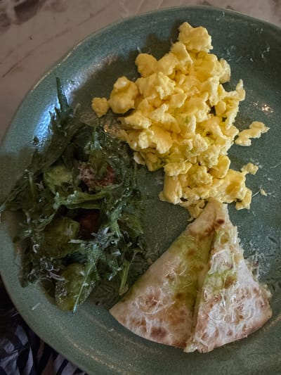 A plate of food consisting of scrambled eggs, a folded quesadilla, and a salad, served in what is presumed to be Ubud, Indonesia. The eggs are pale yellow and fluffy. The quesadilla appears to have a green filling and is sprinkled with cheese. The salad contains green leaves and possible tomato pieces, also topped with cheese. The plate is light green and sits on a surface with a zebra striped pattern, possibly clothing. The lighting is somewhat dim, creating shadows on the food and plate.