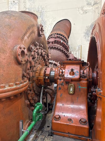 The image displays a close-up of heavy industrial machinery inside a facility located in Pellafol, France. Dominating the scene are several large, aged, reddish-orange metal structures. On the left, a massive, bulbous machine shows extensive oxidation and bears embossed text including "ATELIERS L'ALSTHOM GRENOBLE" and "30 000 GEORGES." This machine features numerous circular bolted flanges and a densely bolted central section. Connected to it by a robust coupling is a smaller, reddish-orange rectangular unit on the right, which has an oval-shaped sight glass or indicator and several wires and small pipes attached. Extending upwards from the central machinery towards the back is a large, reddish-brown, conical and heavily riveted structure. A bright green pipe, connected by two silver flexible corrugated hoses, runs from the lower-left foreground to the base of the left machine. The background consists of light-colored walls displaying significant signs of wear, cracking, and discoloration. The floor beneath the machinery is dark and appears to be a metallic industrial surface. No individuals are present.