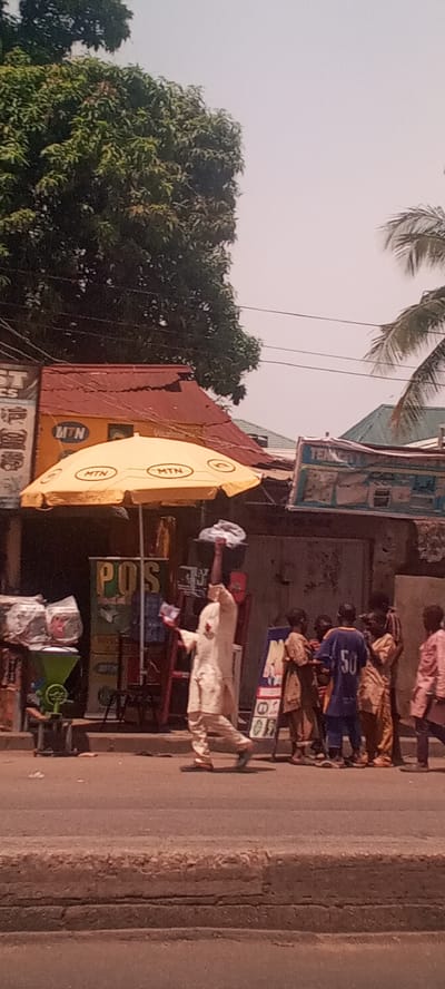 The image captures a bustling street scene in Shaba, Nigeria. A man in a light-colored traditional attire walks with an object balanced on his head, seemingly advertising or selling something with flyers in hand. He is passing by a group of children. Several shops are visible, including one under an MTN-branded umbrella and another bearing the MTN logo on its facade. A POS (Point of Sale) stand is also set up nearby, suggesting commercial activity. The environment includes trees, other buildings, and street-level infrastructure.