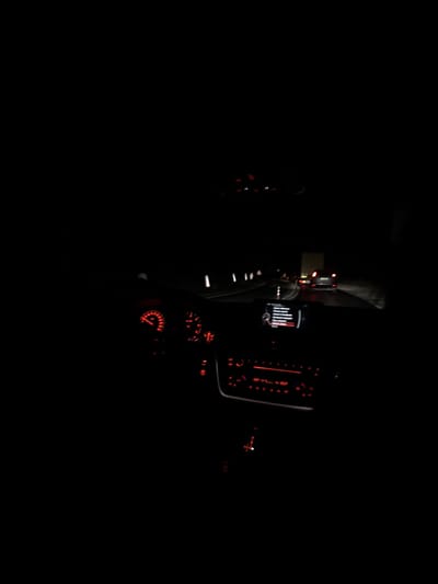 The image depicts the interior of a vehicle at night, viewed from the driver's perspective on a road in Poleto, Bulgaria. The dashboard is illuminated with red lighting, displaying a speedometer on the left and a tachometer on the right. A central infotainment screen shows a menu or text, and below it, climate control buttons are also illuminated in red.
Through the windshield, a dark, wet-appearing road is visible. White reflective road markers line the left side of the thoroughfare, curving slightly to the right. Ahead, multiple vehicles are present, their red taillights distinctly visible against the darkness, indicating motion or a stopped queue. One vehicle, a dark-colored passenger car, is directly in front, while another larger vehicle, possibly a truck, is further ahead on the right. No individuals are discernible within the vehicle or on the road.