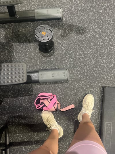 The image shows a person at a gym in Saarbrücken, Germany. The person is wearing white sneakers and light purple athletic shorts. They are standing on a dark gray rubber floor. To their left is a piece of gym equipment with a 10 kg dumbbell attached to it. Next to their foot is a pink and black weightlifting belt.