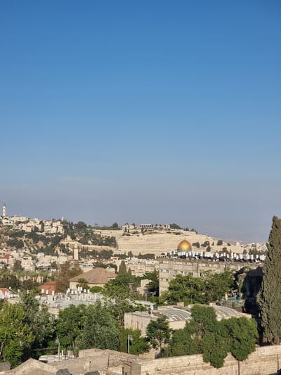 The image shows a daytime view of Jerusalem, Israel, with a clear blue sky occupying the left half. The right half of the image features an urban landscape with buildings, trees, and stone walls. Notably, the Dome of the Rock, a prominent gold-domed structure, is visible in the middle of the scene. The photograph appears to have been taken from an elevated viewpoint, providing a panoramic perspective of the city.