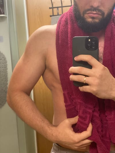 The image shows a close-up, chest-up view of a person holding a smartphone and a burgundy towel in a bathroom. The person, who appears to be male, has a beard and is wearing grey patterned shorts, only partially visible at the bottom of the frame. The person is holding a black smartphone in their right hand, which is positioned over the towel. A thin gold ring is visible on the ring finger of the right hand. The left hand is holding the towel. The background includes a light-colored shower curtain or wall on the left, a wooden door on the right, and a black metal towel rack mounted on the wall. The lighting suggests an indoor environment, likely a bathroom. The image does not contain any elements that would allow for the identification of the location as Koskolovo, Russia.