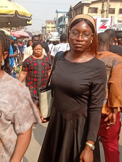 A woman in a black dress and tan headband stands in a crowded outdoor market. She wears glasses and a gold watch. Her bag hangs from her shoulder. People surround her, including a woman in a black and red patterned top. Market stalls with colorful umbrellas are visible in the background, as are buildings and signs. The scene appears busy and lively.