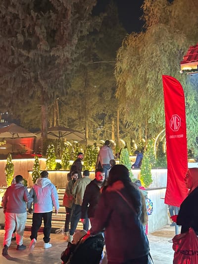 A nighttime outdoor scene features several people walking amongst illuminated Christmas trees. A red MG banner is visible above. Some trees are tall, non-decorated, and dark. The people appear to be in casual clothing and are mostly walking in the same direction. The scene is brightly lit around the Christmas trees and the building behind them.