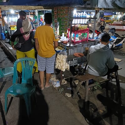 The image shows a street food stall in Kaliwungu, Indonesia (latitude: -6.961, longitude: 110.253). A man in a yellow shirt and striped shorts stands in front of a food cart. He appears to be looking at something in the cart. Behind him, another man in a black jacket and a woman with a headscarf stand near the food cart. The woman is holding a black bag and looking at the man in the yellow shirt. There is a third man sitting on a stool with his back to the camera, wearing a grey hooded sweatshirt and looking down. In the background, there are a few motorcycles and a few people standing or walking. The stall is well-lit, and there are a variety of snacks and drinks for sale. The ground is paved with asphalt.