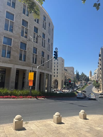The image shows a street scene in Jerusalem, Israel, on a clear day. The sun is high in the sky, indicating it is likely midday. The foreground features a paved sidewalk with decorative stone bollards. To the left, a modern building with a curved facade and stone cladding is visible, featuring numerous square windows. A streetlamp with two ornate fixtures is attached to the side of this building.
The street itself is asphalt, with multiple lanes of traffic. Several cars and vehicles are present, suggesting moderate traffic flow. Along the side of the road, there is a strip of green grass and red flowers. A traffic light is visible, showing a green signal.
In the background, more buildings and trees line the street, leading towards a distant cityscape with some taller structures and what appears to be a clock tower. The overall impression is of an urban environment in daylight.
Visible text on a signpost near the street indicates "No Left Turn to Kariv St." and also provides information about "TRAFFIC CHANGES" in both Hebrew and Arabic. The architectural style of the buildings suggests a contemporary or modern design, characteristic of parts of Jerusalem. There are no people clearly discernible in the image.