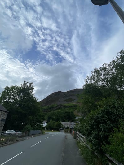 The image captures a view of a road leading into a village nestled at the base of a mountain in Llangynog, United Kingdom. The sky above is filled with a mix of white and grey clouds, with patches of blue visible. The road, paved with asphalt, has white dashed lines indicating lanes. To the left, a stone building and a silver car are visible next to a metal fence. Trees with lush green foliage line the right side of the road, bordering a wooden fence. Further into the scene, several houses with white walls and dark roofs are scattered amongst the trees. The mountain in the background is steep and covered with rocky debris and green vegetation.