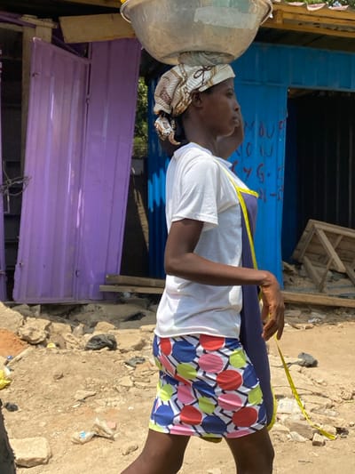 In Ablekuma, Ghana, a woman walks rightward in profile on an uneven, dirt ground under bright daylight. She is carrying a silver-colored metal bowl balanced on a patterned light-colored headscarf. She wears a short-sleeved white t-shirt, a dark blue apron with a yellow strap, and a short skirt with a pattern of multi-colored circles (red, pink, green, yellow, blue) on a white and dark geometric background. Behind her are two corrugated metal structures: a purple one partially open on the left, and a blue one with faint white markings visible on its surface to the right. The ground in the background features rocks, debris, and what appears to be a plastic bottle and a crumpled plastic bag. A wooden structure is visible behind the blue stall.
