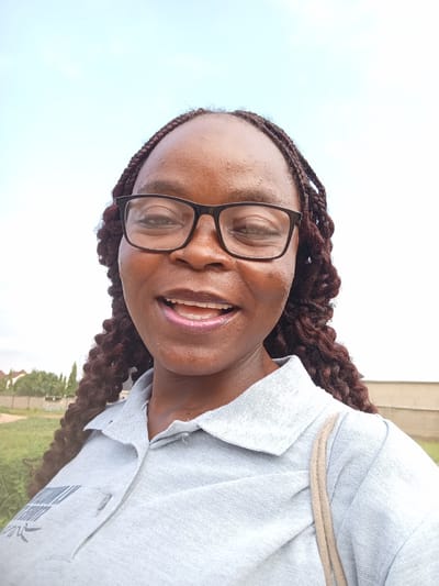 A young African woman wearing glasses and a grey polo shirt smiles at the camera. Her hair is braided with dark brown extensions. She is standing outdoors with a light blue sky in the background. In the distance, there are trees and buildings. The overall scene suggests a casual, outdoor setting in Mayo Dasa, Nigeria.