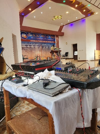 Inside what appears to be a church, a banner reads "EGLISE DU CHRIST AU CONGO" (Church of Christ in Congo), advertising an evangelical assembly. A sound mixing board, along with a laptop and cables, sit on a table covered with white fabric. The interior features a decorated ceiling with a cross design.