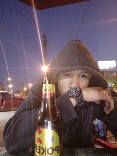 A person in a black hoodie sits at a table with a bottle of Poker beer. The scene is illuminated by a streetlight creating a bright glare. The individual is wearing a large wristwatch. In the background, a red truck is visible, along with other people and buildings in the distance. Power lines crisscross the twilight sky behind a utility pole.