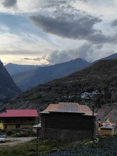 The image depicts a mountainous landscape with a small village nestled in the valley. In the foreground, there are two buildings. The building on the left has a red roof and appears to be a house. To its right, a darker, stone-like building with a weathered, possibly metal roof dominates the center of the frame. Beyond these structures, the terrain slopes upwards, covered with vegetation and dotted with more houses forming the village. Further in the distance, imposing mountains rise, with some peaks snow-capped, under a cloudy sky. The overall scene suggests a rural setting in a mountainous region. Based on the provided context, this scene is located in the city of Gushal, India. No people are visible in the image, and there are no explicit actions or interactions taking place. The environment is characterized by rugged natural terrain and traditional architecture.