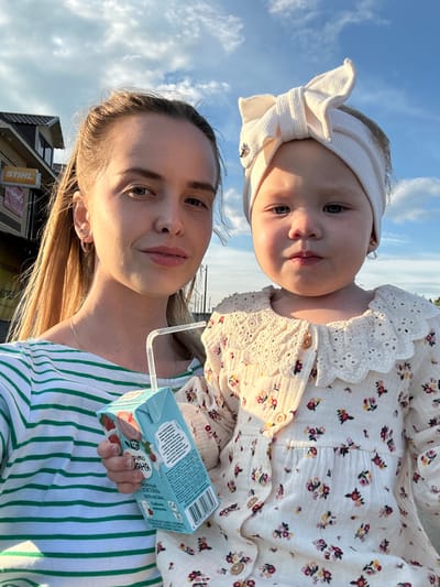 A woman and a young child are standing outside with a building and cloudy sky in the background. The woman is on the left side of the frame, wearing a green and white striped shirt and holding a small juice box with a straw. She has blonde hair pulled back and is looking at the camera. The child is on the right side of the frame, wearing a white floral dress with a large white bow headband. The child is looking at the camera with a neutral expression. The building in the background appears to be a commercial establishment with signage. The lighting suggests it is daytime, likely late afternoon or early evening due to the warm glow. The overall impression is a casual, candid moment captured outdoors. The location is identified as Azinsky, Russia.