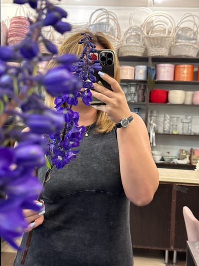 The media file shows a close-up, distorted reflection of a person in a mirror, holding a phone to take a selfie. The person has blonde hair and is wearing a dark, ribbed t-shirt. On their wrist, they wear a black analog watch with a leather strap. The person's right hand, visible in the reflection, has light blue painted fingernails. They are holding a stem of purple flowers, which are also visible in the foreground, slightly out of focus.

In the background, the reflection reveals shelves filled with various items. On the left side of the reflection, there are stacks of small, woven baskets with white handles. To the right of the baskets are shelves containing cylindrical objects wrapped in colorful paper, including orange, pink, and red. Further to the right, more shelves are visible, holding what appear to be glassware or decorative items. The overall setting suggests a shop or a storage area, possibly a craft or flower shop given the presence of floral elements and crafting supplies. The lighting appears to be indoor artificial light. There is no discernible text in the image.