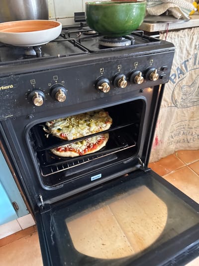 Two pizzas are visible inside a black oven with the door ajar. The pizzas are on oven racks, with the lower pizza covered in red sauce and cheese, and the upper pizza topped with cheese and green olives. The oven is a freestanding gas range with four gold-colored control knobs visible on the front panel. Above the oven, a green bowl sits on a stovetop burner. To the right of the oven, a beige-colored bag with text is leaning against a tiled wall. The floor is covered in orange tiles. The overall scene depicts a kitchen environment. The location is identified as Santa Eulàlia de Ronçana, Spain.