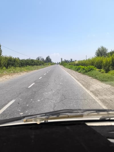 The image shows a view from inside a vehicle looking out at a paved road in Hudayarhan, Uzbekistan. The road has white dashed lines indicating lanes. To the left of the road, there is a grassy verge and then rows of trees. To the right of the road, there is also a grassy verge and dense green vegetation, with more trees further in the distance. The sky is clear and blue with a few faint clouds. There are a couple of distant vehicles on the road. The foreground of the image is dominated by the dashboard and windshield wipers of the vehicle.