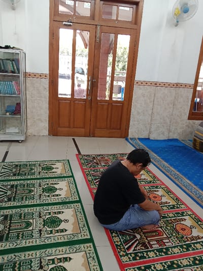 The scene is a mosque in Kecamatan Brangsong, Indonesia, located at coordinates -6.948 latitude and 110.231 longitude. There is one man kneeling on a prayer rug in front of a double wooden door. He is wearing a black t-shirt and blue jeans and has short dark hair. Three green prayer rugs are laid out on the light-colored tiled floor in front of him, extending toward a blue rug against a white wall. There is a bookshelf with books and papers to the left of the door. The walls are tiled and white, with a brown tile trim running along the bottom. A ceiling fan is visible in the upper right corner of the image.