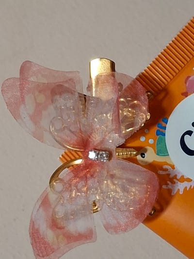 The media file is a close-up, overhead shot of a decorative hair clip. The hair clip is shaped like a pink and white sheer organza bow with gold accents. At the center of the bow, there is a small, clear faceted gem. Attached to the back of the bow is a gold-colored clip mechanism. The bow is fastened to an orange, comb-like object with a decorative pattern. The background is a light, neutral color, suggesting an indoor setting. There are no people or a discernible scene. The lighting is bright, indicating daytime. No text is clearly visible. The overall impression is of a cute and whimsical hair accessory.
