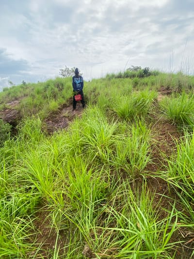 A lone individual stands on a grassy hillside, facing away from the camera. The person wears a dark blue hoodie with a graphic design and dark pants. A bright red bag is held in their hands. The hillside is covered in lush green grass, with some exposed soil and rocks. The sky above is overcast with gray clouds. The location is identified as Achara, Nigeria.