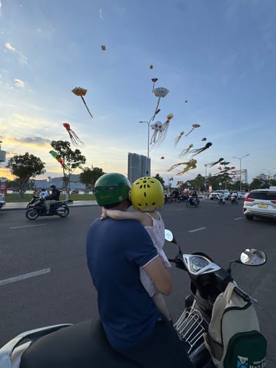 A man and a young child wearing helmets are on a motorcycle, facing away from the camera. The man is wearing a blue shirt, and the child is wearing a pink dress. They are positioned on a paved road with white lane markings. In the background, several other motorcycles and cars are on the road. Above the road, numerous colorful kites are flying in a clear blue sky with scattered clouds. The kites are shaped like various creatures, including jellyfish and a stingray. The scene appears to be set in an urban environment, with buildings visible in the distance. The lighting suggests it is either early morning or late afternoon. The location is Quy Nhon Ward, Vietnam, as indicated by signage on a white SUV in the background.