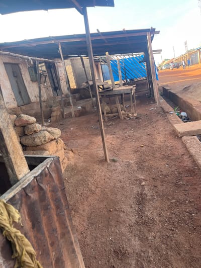 This media file captures an unpaved, reddish-brown earthen area in a rural or semi-urban setting, likely Kuru Dungus, Nigeria, during bright daylight.
On the left side of the frame, several rudimentary structures are visible, primarily constructed from wooden beams and planks. One of these structures features a bright blue tarp or corrugated plastic sheeting as part of its roof or wall. The walls of some buildings appear to be made of light-colored blocks or mud, with partially closed windows and doors, some covered with corrugated metal sheets. Near the base of these structures, piles of rough, light-colored rocks or dried mud bricks are stacked.
The ground across the scene is unpaved red soil, interspersed with small rocks and scattered debris, including plastic items and general refuse, particularly noticeable in a shallow ditch or drainage area.
In the right foreground, a rusty corrugated metal sheet, possibly part of a makeshift roof or barrier, is prominently featured, with a crumpled yellowish-brown piece of fabric draped over it.
In the distant background, an unpaved road runs horizontally. Parked vehicles, including a dark sedan and a lighter-colored car, are visible along this road. Several indistinct human figures, likely three or four, can be seen standing or walking near these vehicles. Utility poles with overhead wires are also present further in the background.
The sky is bright and clear, indicating fair weather and daytime conditions. No specific activity or event is taking place; the scene appears static, depicting an everyday environment. There is no visible text in the image.