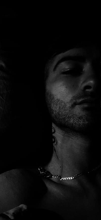 This is a black and white, close-up, vertical, chest-up portrait of a young man with fair skin, a light beard, and dark hair. The man is positioned on the right side of the frame, with the left side mostly in shadow. He is looking downwards and to the left, with his left eye partially visible. He is wearing a silver chain necklace. A tattoo is visible on his neck. The background is dark and nondescript, providing no environmental context. There is no discernible location in Kostenets, Bulgaria, or any other city.
