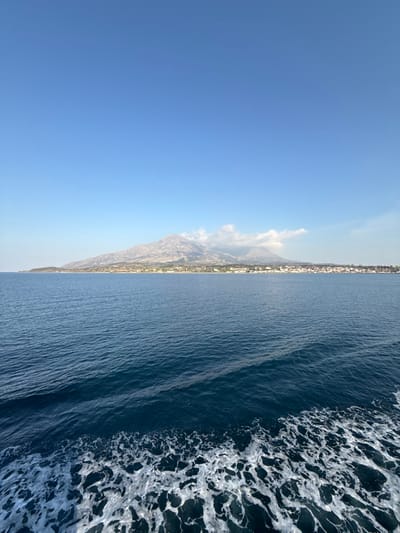 The image presents a panoramic daytime view of the coastline and mountainous interior of Kamariotissa, Greece, observed from a moving vessel on the sea. The foreground is dominated by deep blue ocean water, with a distinct, turbulent white wake indicating forward motion visible in the lower right quadrant. A subtle, linear disturbance extends across the water further into the midground. The midground transitions to a distant landmass featuring a rugged, arid mountain range that rises prominently from the sea. White clouds partially obscure some of the mountain peaks, particularly towards the right side of the frame. Along the base of these mountains, directly at the coastline, a settlement identified as Kamariotissa is visible, characterized by numerous light-colored buildings with scattered reddish roofs. The sky, occupying the upper third of the image, is a clear, bright blue with minimal cloud cover. No individuals or specific actions beyond the implied movement of the viewpoint are discernible.