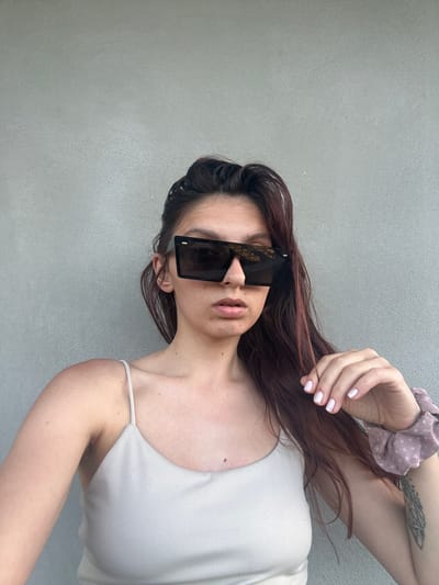 A young woman with dark hair and light skin poses in front of a concrete wall. She is wearing a light-colored tank top and oversized square sunglasses. Her right hand, with light purple nail polish, rests on her chest, near her chin. A light purple polka-dot scrunchie is visible on her left wrist, along with a tattoo on her arm. The background is a plain, light gray concrete wall. The overall lighting suggests an outdoor setting during the day. The location is not discernible from the image alone.