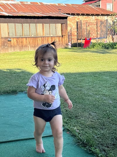 A young child with brown hair and big brown eyes is standing in a grassy backyard. The child is wearing a purple t-shirt with a picture of Minnie Mouse on it and dark blue shorts. The child is looking directly at the camera and smiling. The backyard has a green mat on the left side and grass on the right side. In the background, there is a building with a rusted metal roof and wooden siding. There is also a trampoline and a red slide visible. The scene appears to be in a residential area of Oreshak, Bulgaria.