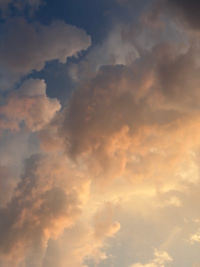 This is a close-up, angled shot of a dramatic sky filled with cumulonimbus clouds. The time of day appears to be late afternoon or early evening, as indicated by the warm, golden light illuminating some of the clouds from the side, creating a striking contrast with the darker, stormier clouds. The clouds exhibit a range of colors, from deep blues and grays in the shadowed areas to soft peaches and yellows where the sunlight catches them. The texture of the clouds is varied, with some appearing fluffy and soft, while others are more dense and textured, suggesting turbulent weather. There are no people or objects visible in the frame, making the sky itself the sole subject. The scene evokes a sense of awe and the power of nature. There is no visible text in the image.