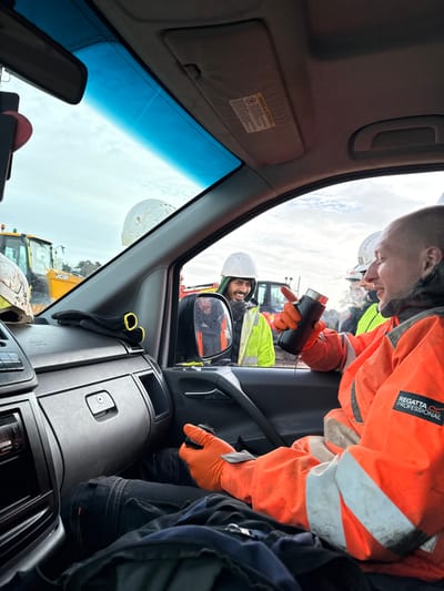 The image, taken from inside a grey vehicle, depicts a construction site in Telford, United Kingdom, under an overcast sky. In the foreground, a bald male, light-skinned individual is seated in the passenger seat, wearing an orange Regatta Professional hi-visibility jacket with white reflective stripes, black trousers, and orange gloves. He holds a black thermal mug with a silver cap in his right hand, from which a dark liquid is visibly spilling onto his jacket. His left gloved hand rests on his leg.
Through the vehicle's open passenger window and reflected in the side mirror, at least two male construction workers are visible outside. One prominent worker, with dark hair and a beard, wearing a white hard hat and a yellow/lime green hi-visibility jacket, smiles and gestures with his right hand while looking into the vehicle. A second male worker, wearing a white hard hat and a hi-visibility jacket, is partially visible to the right of the first. In the background, yellow JCB construction vehicles and what appear to be piles of aggregate are present, confirming an active construction environment. Inside the vehicle, a white hard hat, black gloves, and a dashboard with an audio unit are visible on the dashboard, with a black bag on the floor.
