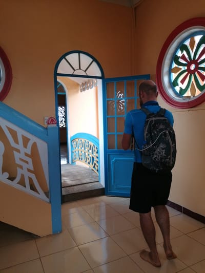 A bald man wearing a blue t-shirt and black shorts, carrying a camouflage backpack, walks barefoot through a hallway. He faces away from the camera, towards a blue doorway. The hallway has orange walls and beige tile floors. To his left, a staircase with a blue railing and white decorative accents is visible. To his right, a circular stained-glass window with red, green, and yellow panes is set into the orange wall. A smaller arched window with a blue frame is visible through the open doorway. The scene appears to be within a building in Lộc Quý, Vietnam, characterized by its colorful architecture and decorative elements.