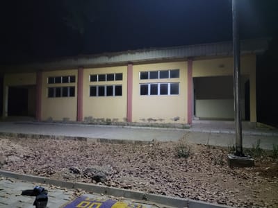 A yellow building with a symmetrical facade sits beneath a dark sky, illuminated by unseen light sources. Three sets of windows punctuate the walls, interspersed with reddish-brown vertical sections. Open doorways flank the windowed sections. The ground in front of the building is uneven, covered in debris and leaves. A pole stands near the right side of the structure. A pair of sandals rest on a striped mat at the bottom left. The entire scene is captured in a dimly lit environment.